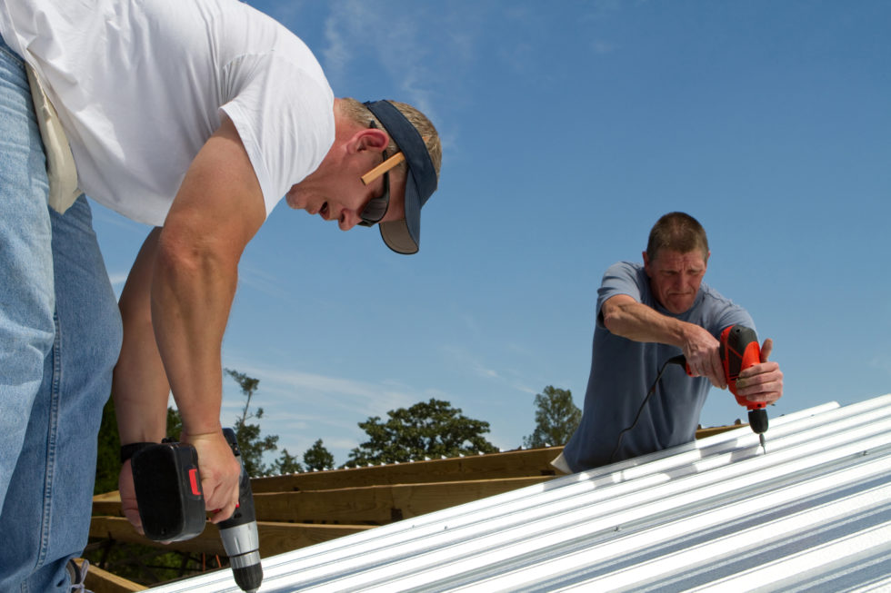 Construction roofing crew uses power tools to screw and fasten sheet metal to the roof rafters of a building to show why proper installation is important for how long does metal roofing last.